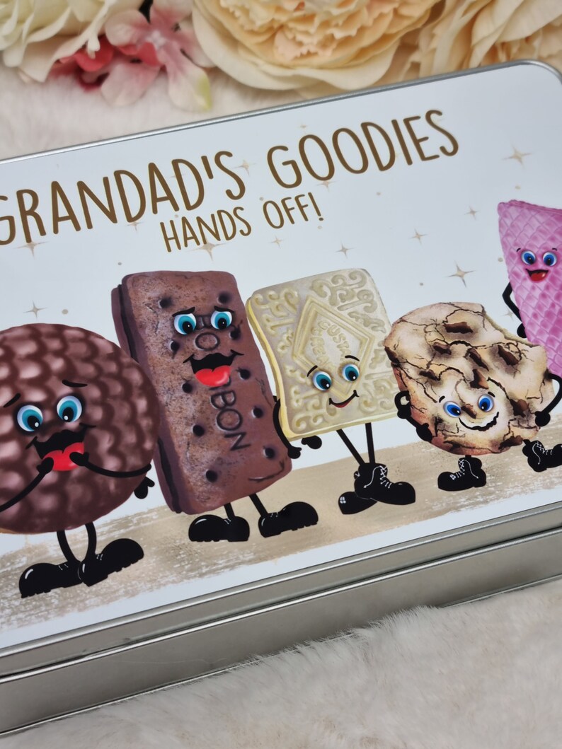 Puede incluir: Una lata de plata con fondo blanco y el texto "Grandad's Goodies Hands Off!" en letras marrones. La lata presenta personajes de dibujos animados de varias galletas con caras, brazos y piernas.