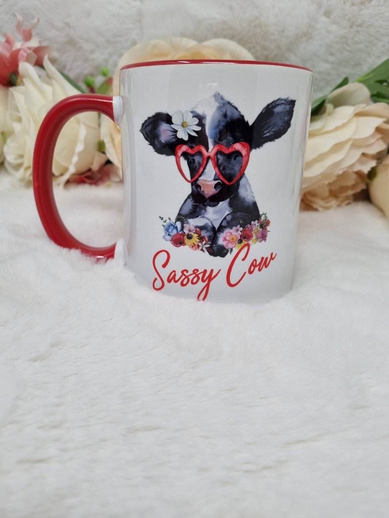 May include: White ceramic mug with a red rim and handle. The mug features a cartoon illustration of a black and white cow wearing red heart-shaped sunglasses and a flower in its hair. The text "Sassy Cow" is printed in red below the cow.