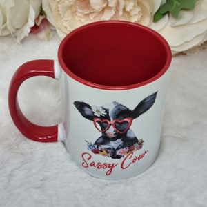 May include: White ceramic mug with a red handle and rim. The mug features a cartoon illustration of a black and white cow wearing heart-shaped sunglasses and a flower crown. The text "Sassy Cow" is printed in red below the cow.