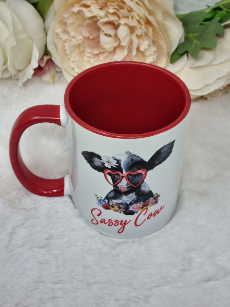 May include: White ceramic mug with a red handle and rim. The mug features a cartoon illustration of a black and white cow wearing heart-shaped sunglasses and a flower crown. The text "Sassy Cow" is printed in red below the cow.