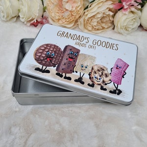 Puede incluir: Una lata de metal plateada con una tapa blanca que presenta una ilustraci&oacute;n de dibujos animados de varias galletas con caras. La tapa dice "Grandad's Goodies Hands Off!"