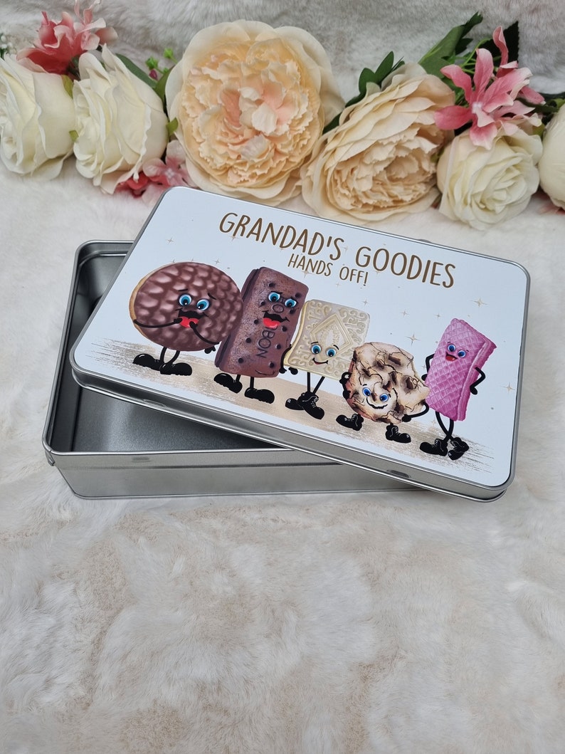 Puede incluir: Una lata de metal plateada con una tapa blanca que presenta una ilustraci&oacute;n de dibujos animados de varias galletas con caras. La tapa dice "Grandad's Goodies Hands Off!"