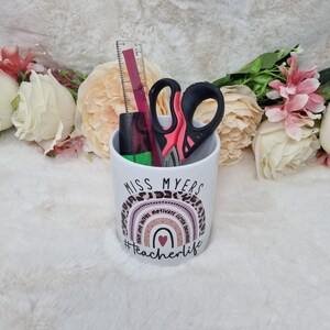 May include: White ceramic pencil holder with a rainbow design and the text "Miss Myers #teacherlife". The rainbow has the words "teach love write motivate listen believe" around it. The holder has a ruler, scissors, and a black marker inside.