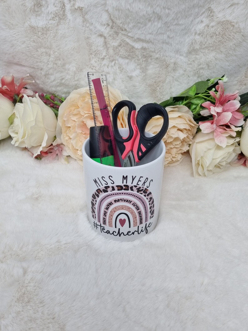 May include: White ceramic pencil holder with a rainbow design and the text "Miss Myers #teacherlife". The rainbow has the words "teach love write motivate listen believe" around it. The holder has a ruler, scissors, and a black marker inside.