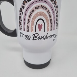 May include: White travel mug with a pink, brown, and black rainbow design. The rainbow has the words "Teach Love Inspire Motivate Listen Encourage" around it. The mug also has the text "Miss Borsberry" in black cursive writing.