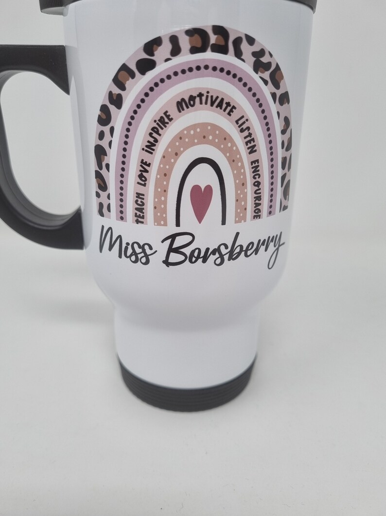 May include: White travel mug with a pink, brown, and black rainbow design. The rainbow has the words "Teach Love Inspire Motivate Listen Encourage" around it. The mug also has the text "Miss Borsberry" in black cursive writing.