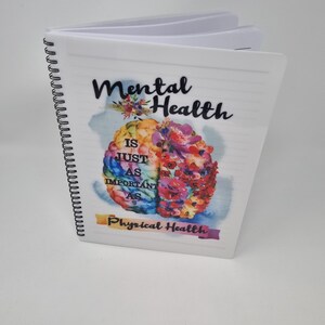 K&ouml;nnte beinhalten: Ein wei&szlig;es Spiralnotizbuch mit dem Text "Mental Health is just as important as Physical Health" auf dem Cover. Der Text ist schwarz und das Cover zeigt eine farbenfrohe Aquarellillustration eines Gehirns mit Blumen.
