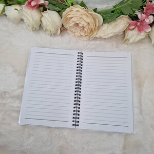 K&ouml;nnte beinhalten: Ein wei&szlig;es Spiralblock mit linierten Seiten. Der Block ist ge&ouml;ffnet und liegt flach auf einer wei&szlig;en, flauschigen Oberfl&auml;che. Eine Girlande aus wei&szlig;en und rosa Blumen befindet sich &uuml;ber dem Block.