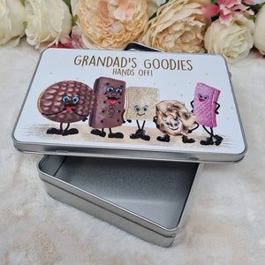 Puede incluir: Una lata de metal plateada con una tapa blanca que presenta una ilustraci&oacute;n de dibujos animados de varias galletas con caras. La tapa tiene el texto "Grandad's Goodies Hands Off!" impreso en ella.