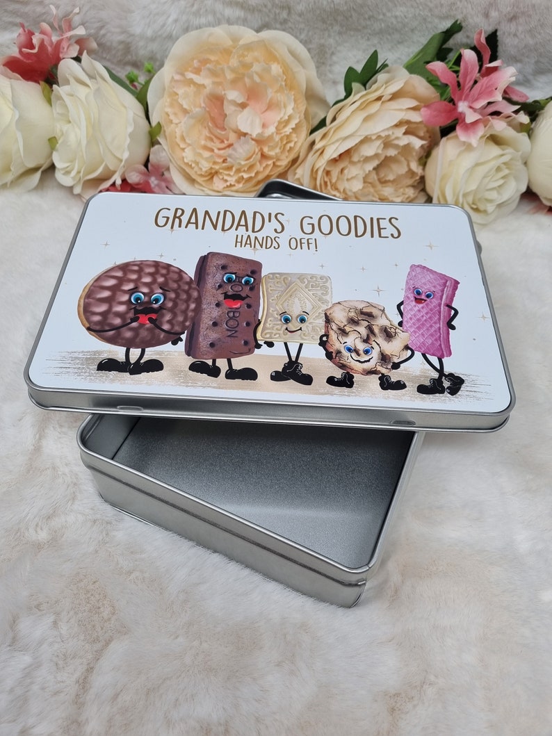 Puede incluir: Una lata de metal plateada con una tapa blanca que presenta una ilustraci&oacute;n de dibujos animados de varias galletas con caras. La tapa tiene el texto "Grandad's Goodies Hands Off!" impreso en ella.