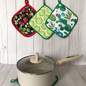 May include: Three colourful pot holders with different patterns: strawberries, limes and cacti. The pot holders are made of fabric and have a green border. A beige pot with a glass lid is sitting on a green pot holder.