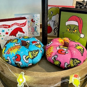 May include: Two decorative Grinch-themed pin cushions in the shape of pumpkins. One is blue with Grinch and Santa Claus graphics, the other is pink. The background includes Grinch-themed holiday decor.