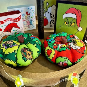 May include: Two fabric pumpkins decorated with a green, red, and yellow Grinch-themed pattern. The pumpkins are sitting on a wooden tiered tray with a string of white lights.