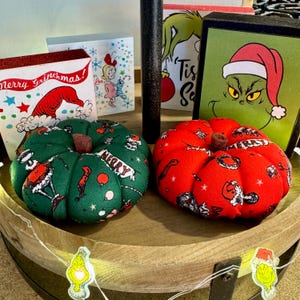 May include: Two fabric pumpkins decorated with a Christmas theme. One pumpkin is green with a red and white "Merry" banner and the other is red with white stars. The pumpkins are sitting on a wooden tiered tray with Christmas decorations.