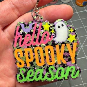 May include: A keychain featuring a multi-colored glitter background with the text hello SPOOKY season in pink, orange, and green. The design includes a small white ghost, a purple bat, and several yellow stars embedded within the resin surface.