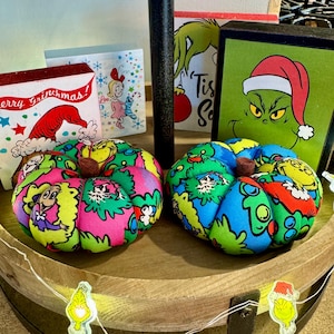May include: Two handmade fabric pumpkins decorated with characters from the Grinch and the Cat in the Hat. The pumpkins are green, pink, blue, and red with a variety of characters and designs.