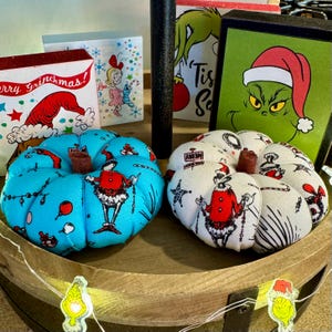 May include: Two handmade fabric pumpkins decorated with a Grinch theme. One pumpkin is blue with white and red accents, and the other is white with red and black accents. Both pumpkins feature a Grinch character holding a string of lights.