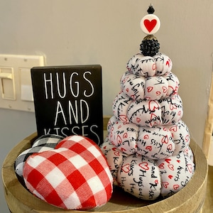 Könnte beinhalten: Ein mehrstufiger Stapel aus Stoffherzen mit dem Aufdruck "Hugs and Kisses". Die Herzen sind weiß mit roten Herzen und Schriftzügen. Der Stapel ist mit einem roten Herzen und einer schwarzen Perle gekrönt. Der Stapel steht auf einem Holztablett mit einem schwarzen Schild, auf dem "Hugs and Kisses" steht, und einem rot-weiß karierten Herzen.