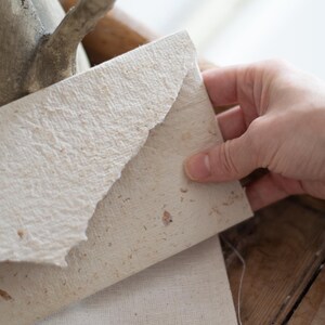May include: A white envelope made of textured paper with a rough, handmade look. The envelope is folded in half and has a natural, rustic feel.