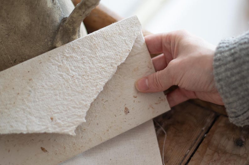 May include: A white envelope made of textured paper with a rough, handmade look. The envelope is folded in half and has a natural, rustic feel.