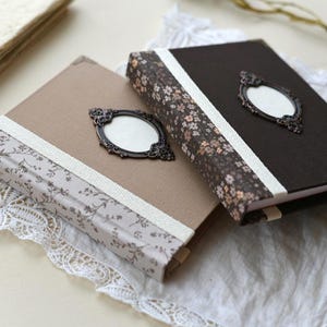 May include: Two decorative journals with floral patterned spines and ornate metal frames. One journal is beige, the other is dark brown. Both have white ribbon accents and are resting on a white lace cloth.