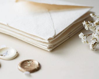 May include: A stack of off-white envelopes with textured edges, accompanied by three circular wax seals in cream and metallic gold tones, and a small sprig of dried white baby's breath flowers arranged to the side of the stationery set.