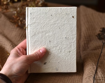 Puede incluir: Un cuaderno rectangular con tapa dura hecha de papel artesanal texturizado en color blanco roto. La cubierta presenta motas y fibras orgánicas visibles incrustadas en la pulpa del papel, creando una textura rústica e irregular en la superficie.