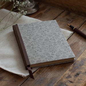 May include: A closed journal with a floral pattern cover and a brown spine and bookmark. A brown pen with gold accents rests on a wooden surface next to the journal. The journal is on a white cloth.