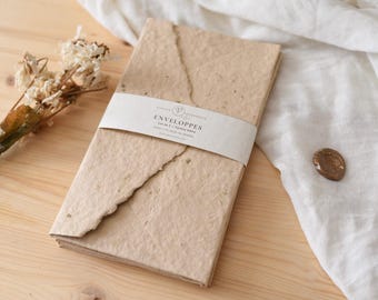 May include: A stack of handmade paper envelopes, wrapped with a white band that reads "ENVELOPPES". The envelopes are a light brown color with a textured, torn edge. Dried flowers and a wax seal are also visible.
