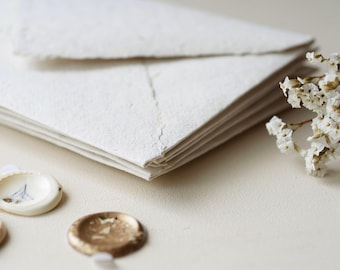May include: A stack of off-white handmade paper envelopes with deckled edges, accompanied by three small circular wax seals in cream, gold, and metallic tones, and a small sprig of dried white flowers positioned to the right of the envelopes.
