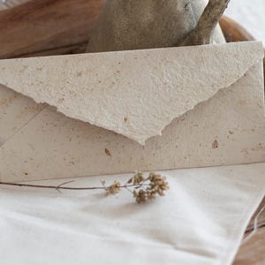 May include: A white envelope made of recycled paper with a textured surface. The envelope is lying on a white cloth surface.