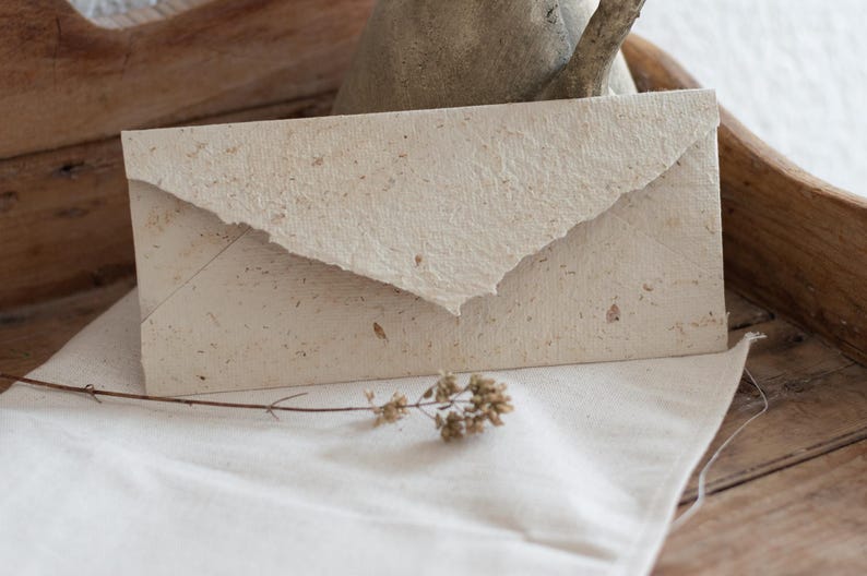 May include: A white envelope made of recycled paper with a textured surface. The envelope is lying on a white cloth surface.