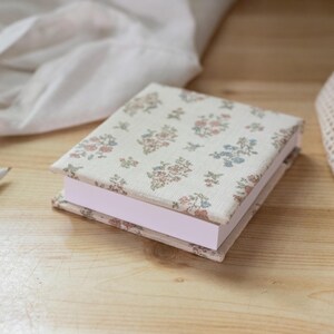 May include: A square notepad with a fabric cover featuring a repeating floral pattern in muted pink, blue, and green tones on an off-white background. The notepad has a thick stack of plain white paper sheets and a sturdy, matching fabric-wrapped base.
