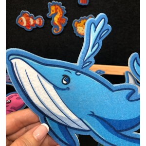 May include: A close-up of a blue whale-shaped felt cutout with a white belly and a smiling face. Other felt sea creatures, including a seahorse, fish, and a pufferfish, are visible in the background. The cutouts are likely for educational or decorative purposes.