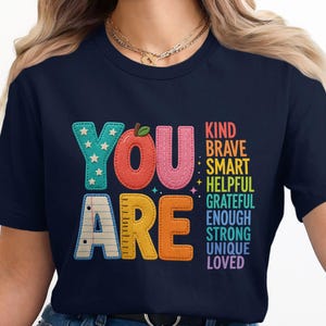 Puede incluir: Una camiseta azul marino con el texto YOU ARE en letras grandes y coloridas con estilo de costura. La palabra YOU es de color turquesa, rojo y rosa, mientras que ARE es blanca, amarilla y naranja. A la derecha, las palabras KIND, BRAVE, SMART, HELPFUL, GRATEFUL, ENOUGH, STRONG, UNIQUE, LOVED están impresas en una lista vertical.