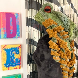 May include: A handmade crochet frog hat in shades of green, brown, and tan, displayed on a black mannequin. The hat features large, round eyes and a textured design. The hat is a whimsical accessory.