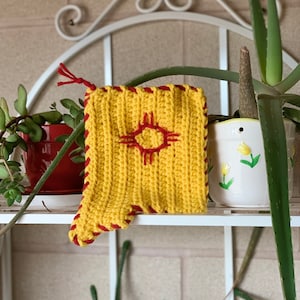 Könnte beinhalten: Ein gelber Häkel-Topflappen mit einem roten Häkel-Symbol in der Mitte. Der Topflappen hat einen roten Häkel-Rand.