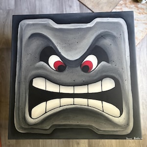 May include: A square, grey, cartoon Thwomp face with an angry expression. The Thwomp has red eyes, a wide open mouth with white teeth, and a dark grey border. The artwork is signed "Danny Koehler '19".