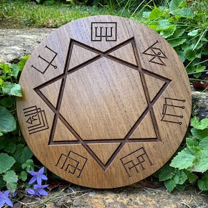 May include: A circular wooden plaque featuring a carved star and geometric symbols. The wood has a warm brown hue, with deeply etched symbols. The plaque is displayed outdoors amidst greenery and small purple flowers.