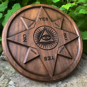 Może przedstawiać: Drewniana plansza Ouija z gwiaździstym wzorem i napisami "YES", "NO", "MAYBE", "UNKNOWN", "NEVER" i "ALWAYS" wygrawerowanymi wokół obwodu. W centrum planszy znajduje się symbol wszechwidzącego oka.