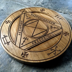 Op de afbeelding: Een houten cirkel met een gesneden ontwerp met een driehoek met de woorden "AKTIOPHIS", "ERESHKIGAL" en "NEBOTOSCOALETH" eromheen gegraveerd.