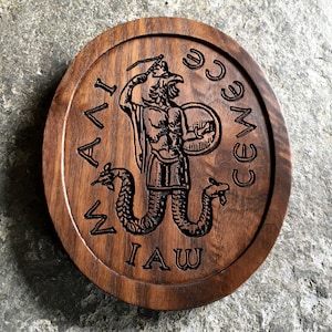 May include: A round wooden plaque with a carved image of a winged, serpent-tailed figure holding a sword and shield. The plaque is inscribed with the words "MAIAI CEWCE IAW".