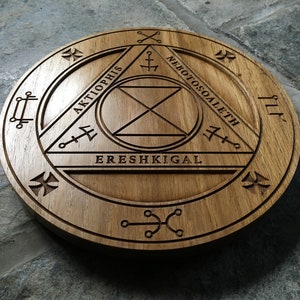 Op de afbeelding: Een houten cirkel met een gesneden ontwerp met een driehoek met de woorden "Aktophis", "Ereshkigal" en "Nebotoscoalethe" eromheen gegraveerd.