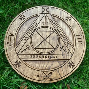 Op de afbeelding: Een houten cirkel met een driehoek erin gegraveerd. De driehoek heeft de woorden "Aktiophis", "Nebotossoaleth" en "Ereshkigal" eromheen geschreven. De cirkel heeft ook andere symbolen en markeringen.