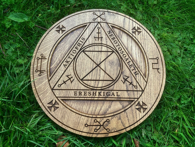 Op de afbeelding: Een houten cirkel met een driehoek erin gegraveerd. De driehoek heeft de woorden "Aktiophis", "Nebotossoaleth" en "Ereshkigal" eromheen geschreven. De cirkel heeft ook andere symbolen en markeringen.