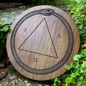 Op de afbeelding: Ronde houten plaat met een donkerbruine afwerking. De plaat heeft een gedetailleerde gravure van een ouroboros slang die een driehoek met de letters I, A en Ω omsluit. De tekst "THE HERMETIC WORKSHOP+" is zichtbaar.