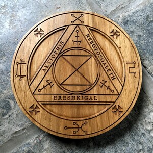 Op de afbeelding: Een houten cirkel met een geometrisch ontwerp en de woorden "Aktiophis", "EreShkigal" en "Nebotosoaleth" die rond de omtrek zijn gegraveerd.