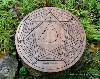 May include: A round, wooden decorative piece with intricate geometric designs and Hebrew text etched into its surface. The design includes triangles, circles, and intersecting lines, with a dark brown finish. The piece is set against a backdrop of green grass and foliage.