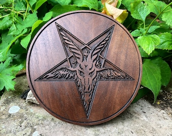 Op de afbeelding: Een ronde houten plaat van donkerbruin hout met een gegraveerd pentagram met een geitenkop in het midden.