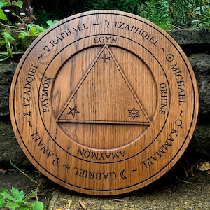 Pode incluir: Um círculo de madeira com um triângulo gravado e os nomes de anjos ao redor da borda. Os nomes incluem Rafael, Tzadkiel, Miguel e Kamael.